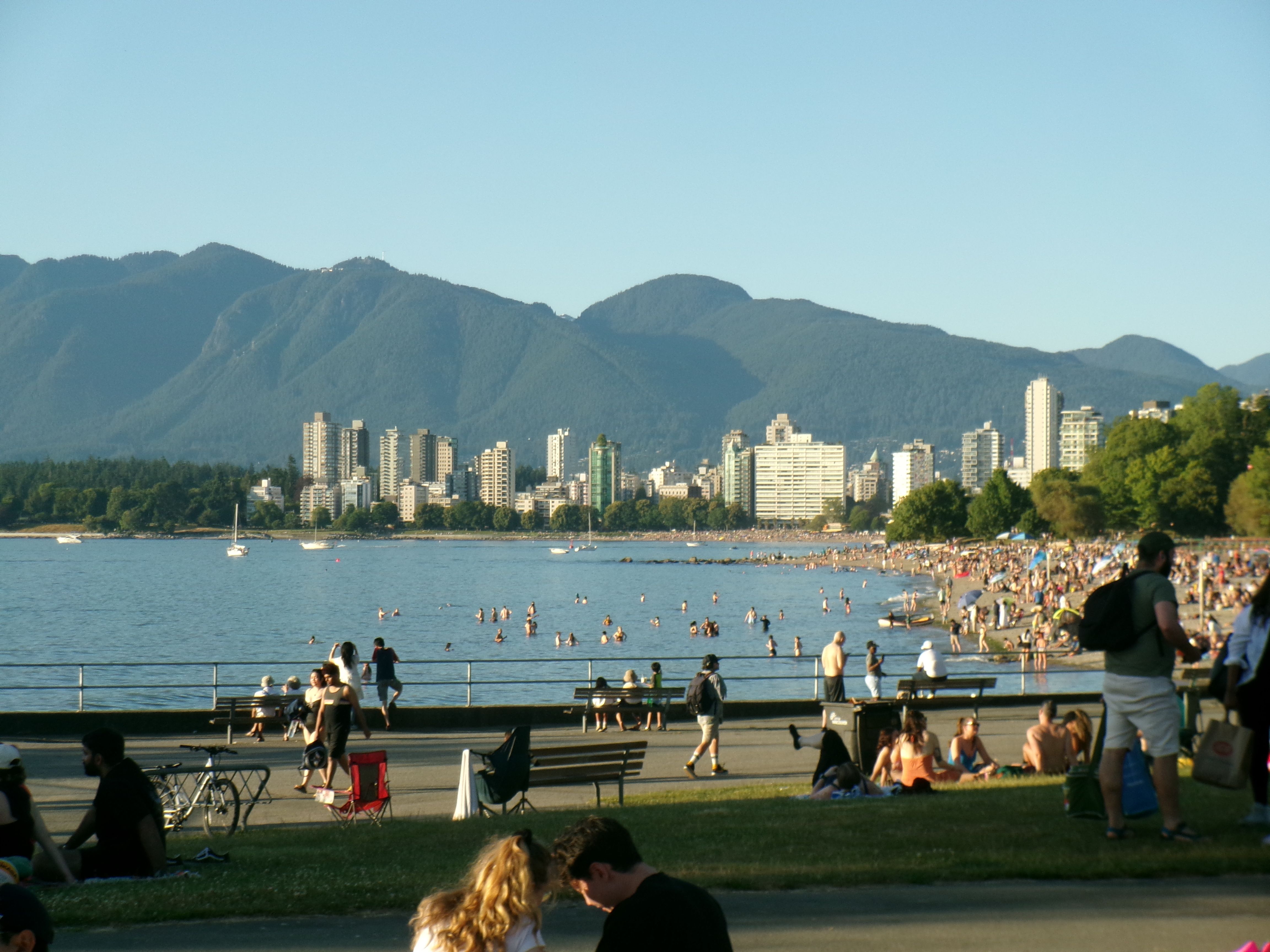 People beside a waterfront with buildings and mountains beyond