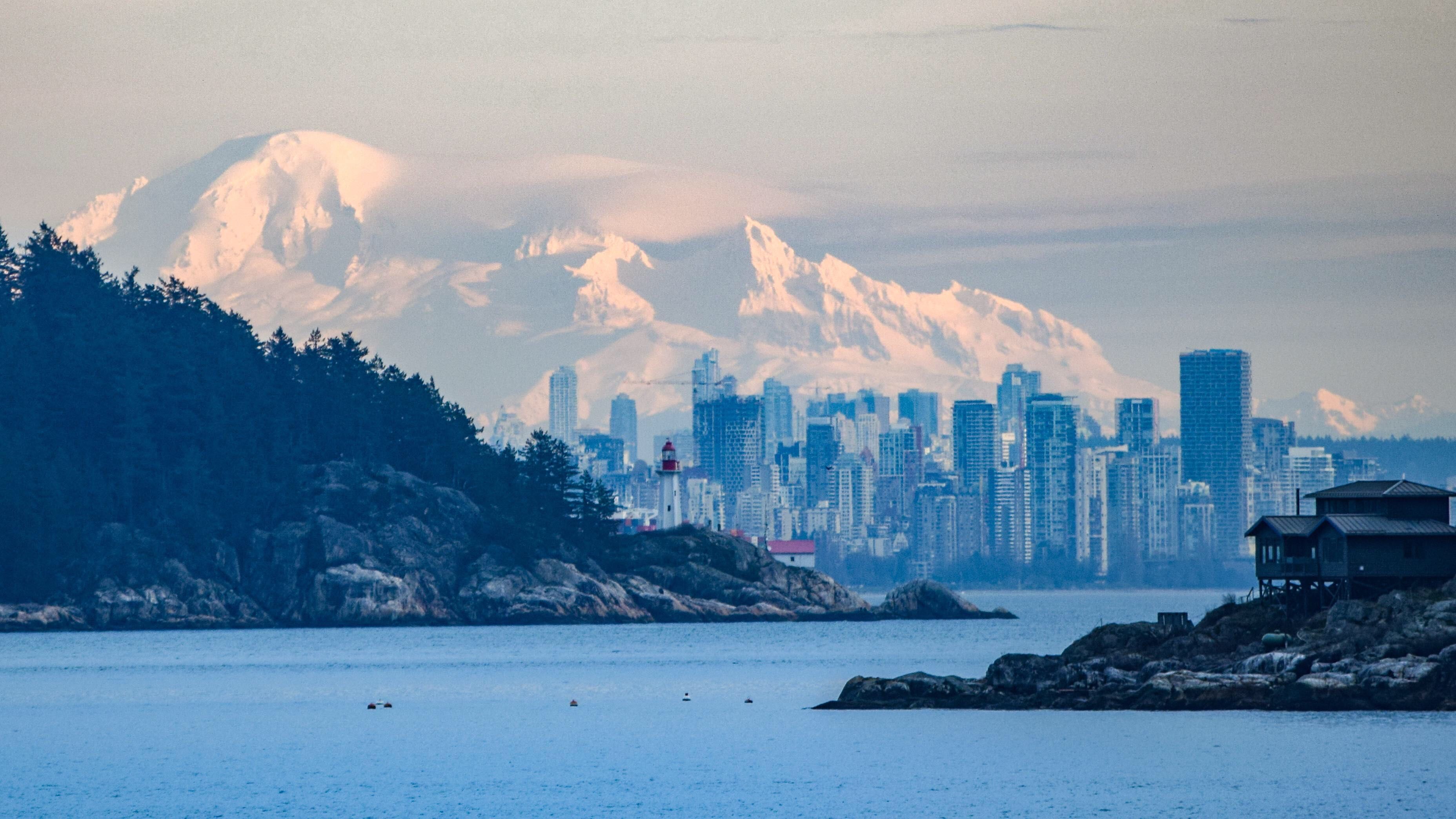 Coastal water, wooded shoreline and a city skyline beneath snowy mountains
